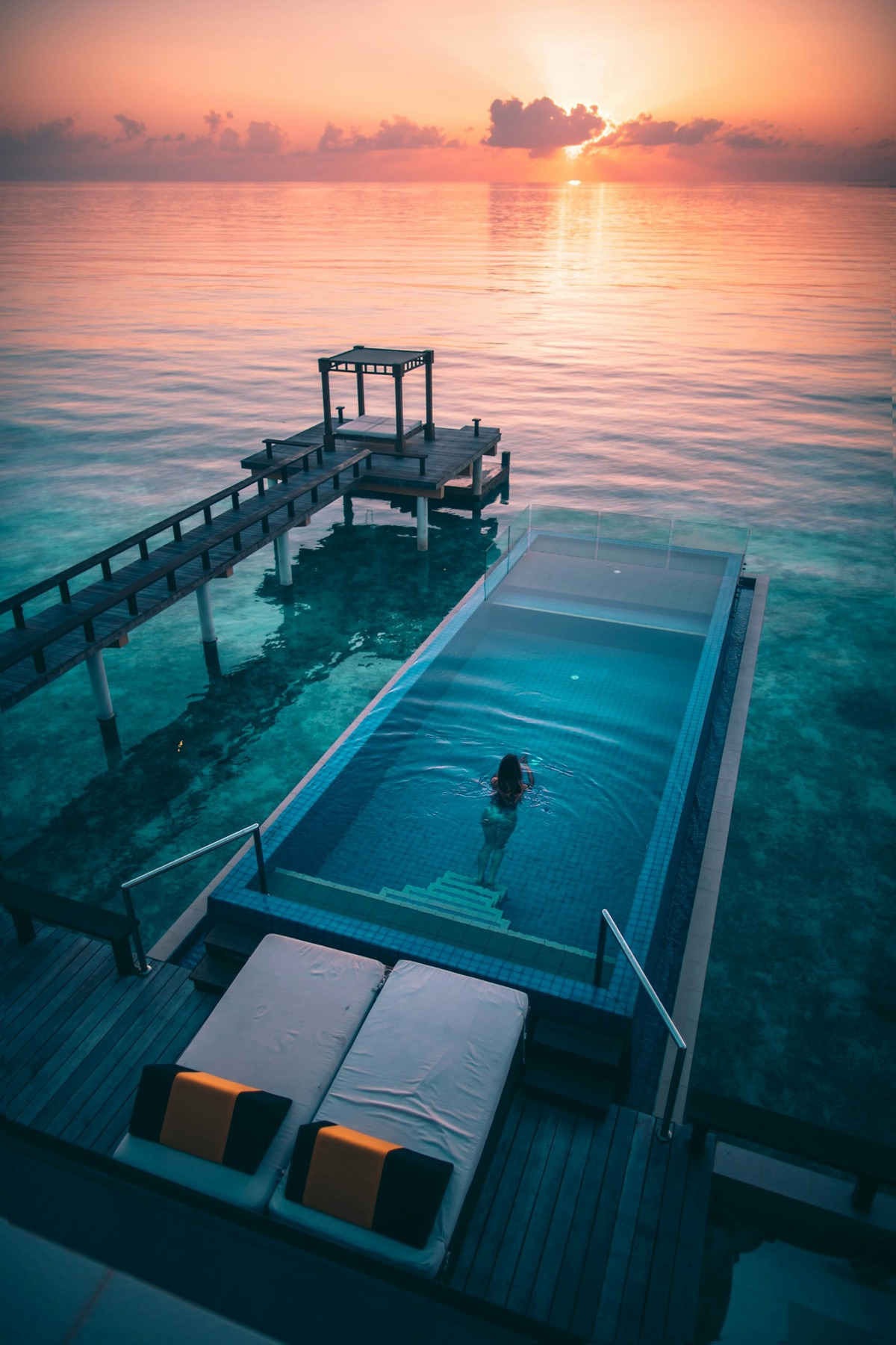 An overwater infinity pool at sunset with a swimmer and daybeds.