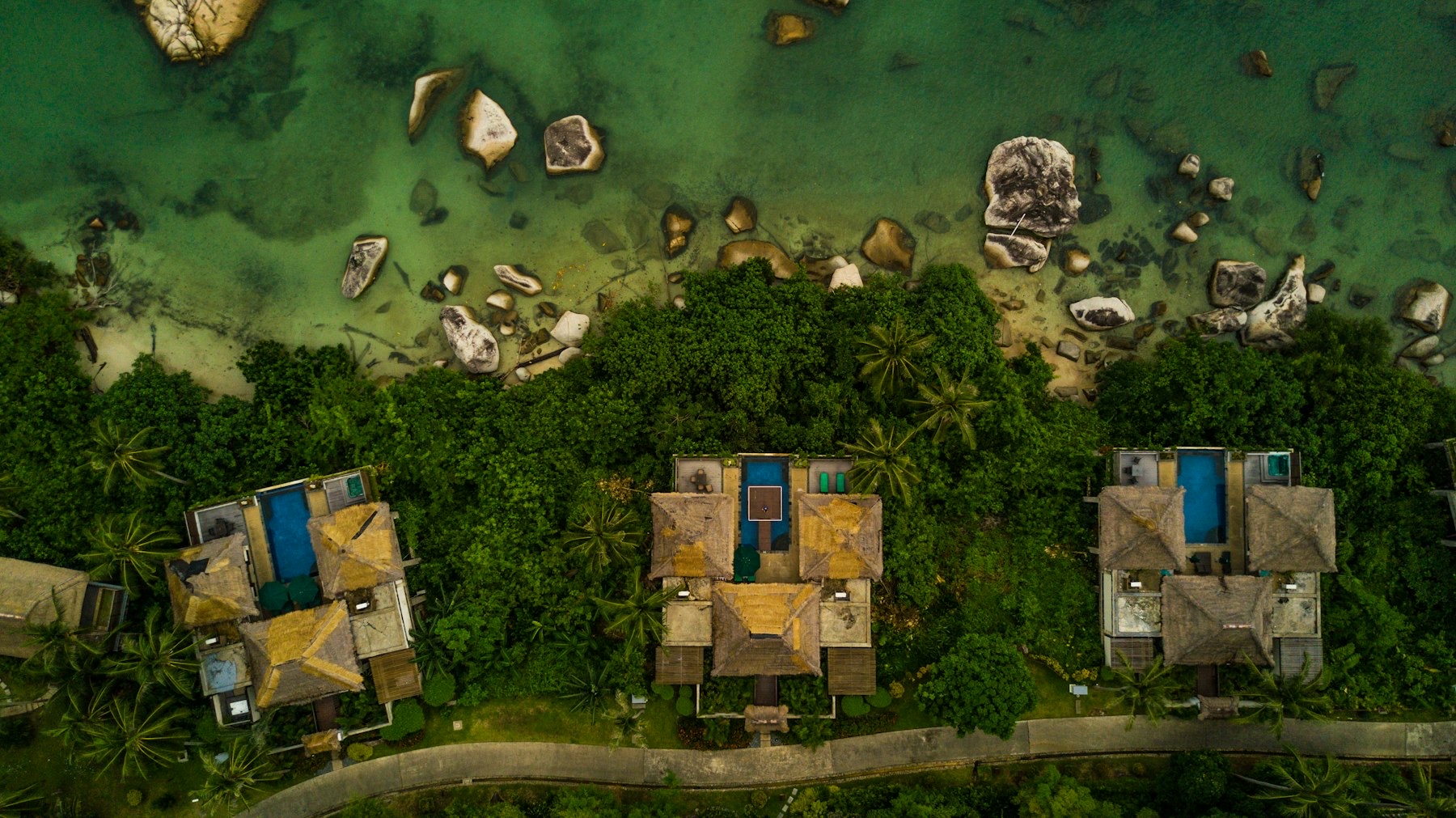 Drone view of private pool villas set in jungle canopy beside the sea.